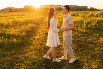 Happy young couple walking toward each other, hugging and kissing on beautiful green meadow in summer evening during golden sunset with warm sunlight. Romantic man and woman enjoying time outdoors.
