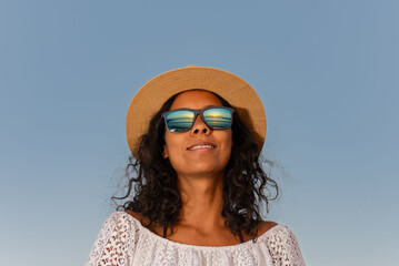 Beautiful brunette Latina woman smiling wearing a hat and white dress with the summer sunrise reflected in her sunglasses