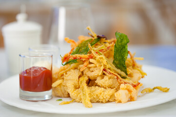 Close-up of calamari rings with fried rucola and chili sauce. Battered squid appetizer on a white plate. Seafood meal on a cafe table. Horizontal food background. Restaurant menu concept.