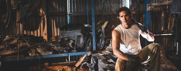 Young and handsome muscular and strop scrap material man in sleeveless shirt sitting on material in junkyard while looking away while taking rest and break from work during in warehouse