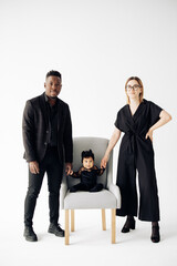 Happy interracial family stands near their baby daughter sitting in armchair.
