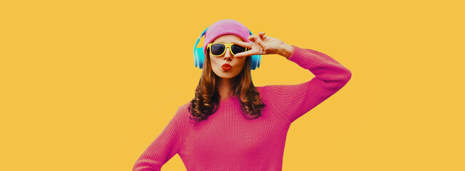 Portrait of modern young woman in wireless headphones listening to music wearing knitted sweater, pink hat on yellow background
