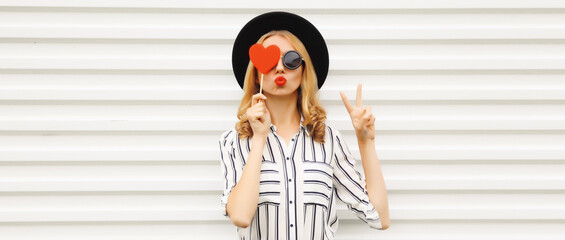 Portrait of beautiful young woman covering her eyes with red heart shaped lollipop wearing black round hat on white background