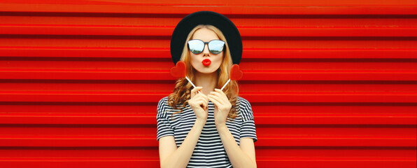 Portrait of beautiful young woman with red heart shaped lollipop blowing her lips with lipstick...