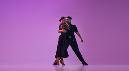 Young talented man and woman, professional dancers in stylish costumes performing tango on purple background on neon lights. Concept of , lifestyle, action, beauty of movements, emotions, fashion, art