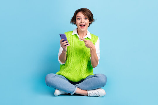 Full size photo of positive girl with brown hairdo wear green vest jeans impressed directing at phone isolated on blue color background
