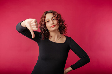 Young redhead woman wearing black ribbed dress isolated over red background not believing the lies, looks at the camera and gives the thumbs down sign. Don't tell lies. I didn't believe it.