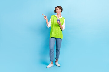 Fototapeta na wymiar Full size photo of cute girl with brown hairdo wear green vest staring indicating empty space hold phone isolated on blue color background
