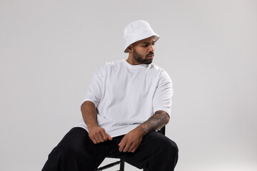 Attractive african american man with a beard dressed in a white t-shirt and panama sits on a chair.