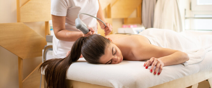 Extracorporeal shockwave therapy in beauty salon. Physical therapy for neck and back muscles,spine with shock waves. Young woman wearing protective mask during treatment in salon.