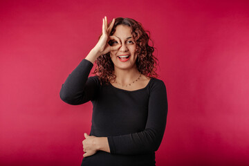 Happy woman wearing black dress standing isolated over red background looking through okey sign. Happy, confident and everything is fine. Face expressions, emotions, and body language.