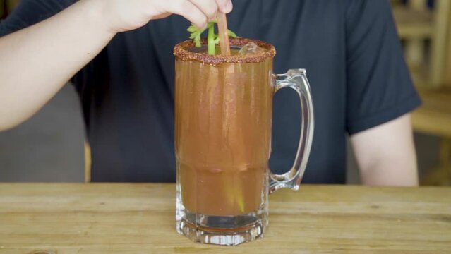 Hombre bebiendo una deliciosa michelada preparada con pepino, camar&oacute;n, chile chamoy, apio y sangrita sobre mesa de madera.