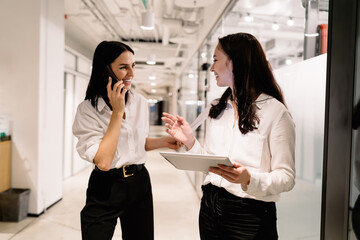 Smiling woman with tablet talking to cheerful colleague using smartphone