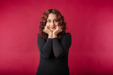 Young beautiful woman wearing black ribbed dress isolated over red background hands in fists, touching chin, looks excitedly at the camera and is dumbfounded. Can't wait to share what has heard.