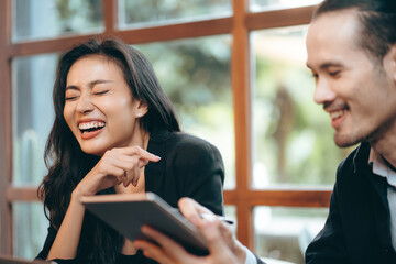 Smiling and laughing young Asian successful business woman discussing work with male colleague and partner in modern office while using digital tablet