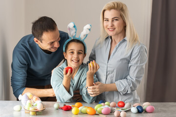 Naklejka premium Joyful family wearing bunny ears headbands gathering at table in modern light kitchen and paining Easter eggs together.