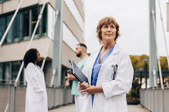 Mature female doctor looking away while holding clipboard and book outside hospital