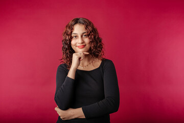 Fototapeta premium Beautiful redhead woman wearing black dress standing isolated over red background touches chin and looks suspiciously at the camera. Trying to find ideas.