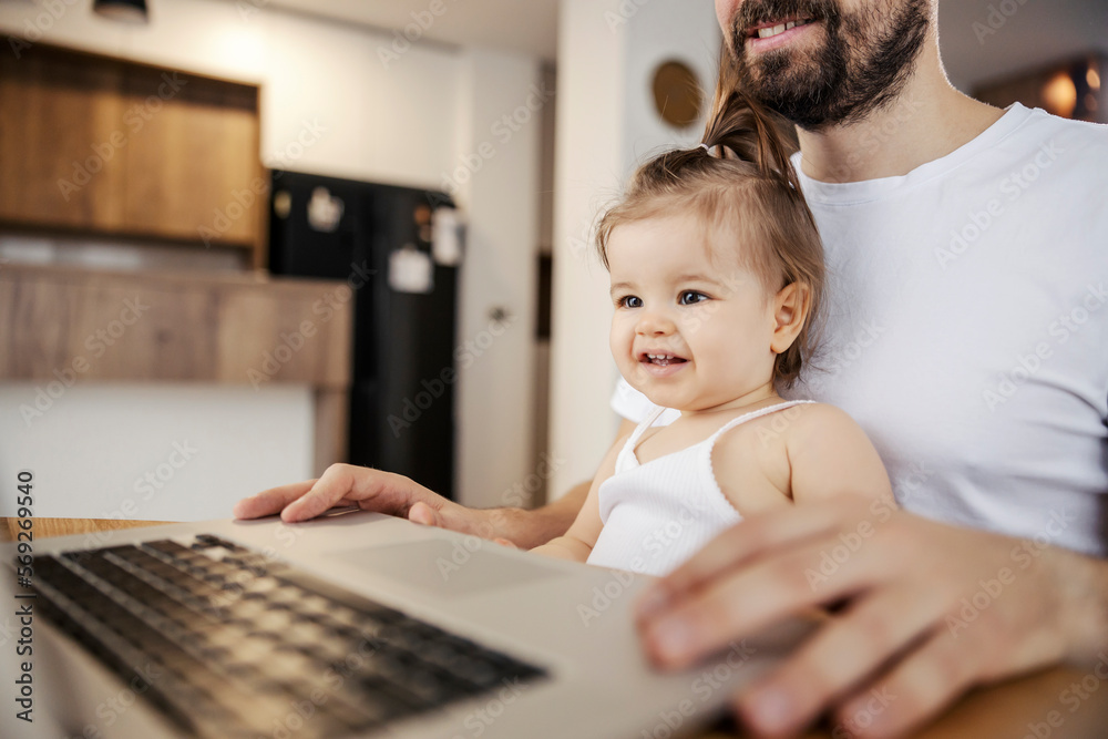 Wall mural portrait of a happy little girl sitting in her father's lap and watching while he works on a laptop.