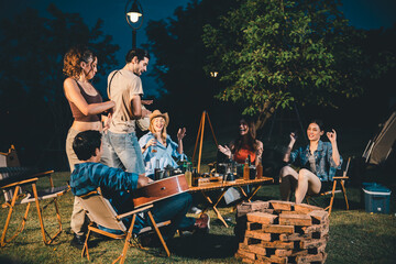 Group of young male and female friends enjoying under night sky near lake while partying and drinking beer playing musical instrument guitar and singing songs