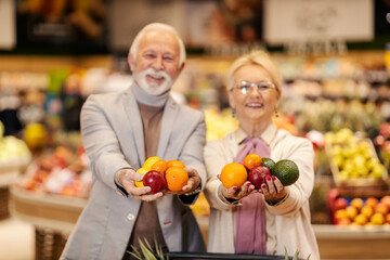 A healthy old couple is showing fresh fruits at the camera while standing at the supermarket.