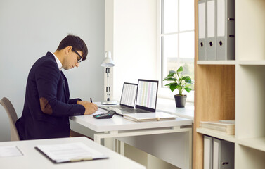 Busy financial accountant working in modern light office interior. Young man in suit and glasses sitting at desk with two laptop computers by window, using spreadsheets and taking notes on paper