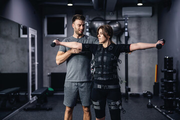 Fototapeta na wymiar Electrical muscle stimulation training concept and EMS suit. Male coach in grey sportswear helping and showing woman in EMS suit the exercise with dumbbells indoor gym with equipments