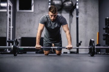Obraz na płótnie Canvas Focus on training and concentration on exercise. A young attractive man in a gray T-shirt rests his hands on a barbell and squats in the gym. Initial body posture and sports discipline