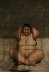 plus size filipino woman with a shaved head hands on head smiling sitting cross legged on the floor