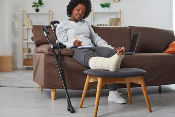 Fototapeta na wymiar Attractive smiling african american woman works at home in the living room with laptop and makes notes in notebook, sitting on the sofa with broken bandaged leg lying on stool. Crutches are nearby.