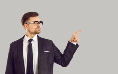 Confident handsome young businessman pointing finger at content or copy space on gray background. Man in black suit, glasses and with tie recommending something or advertising. Business concept.