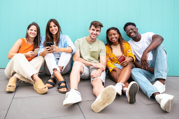 multiethnic group of cheerful friends together outdoors with mobile phone sitting on the floor and looking at camera - education concept