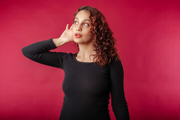 Beautiful redhead woman wearing black dress standing isolated over red background bewildered,...