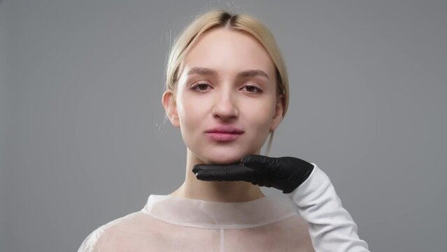 The hands of a cosmetologist surgeon in black gloves touch a woman's face isolated on a light background, a beautiful girl with clean skin without makeup checks the skin of the face at a cosmetologist