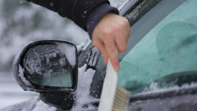 Unrecognizable man with brush for cleaning snow from car side window shield outdoors on heavy snowfall, travel preparation close up. Snowstorm in winter season. Vehicle care in blizzard