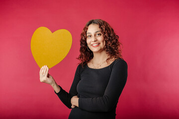 Cute caucasian woman wearing black ribbed dress isolated over red background poses smiling with a cardboard heart shape. Looking at the camera, at you, at lover. It could be a gift.