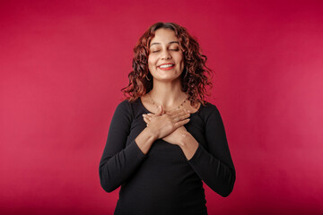 Cheerful redhead woman wearing ribbed dress isolated over red background hold hands on heart chest feel grateful and thankful. Show gratitude, love and care, pray or visualize. Religion, faith concept