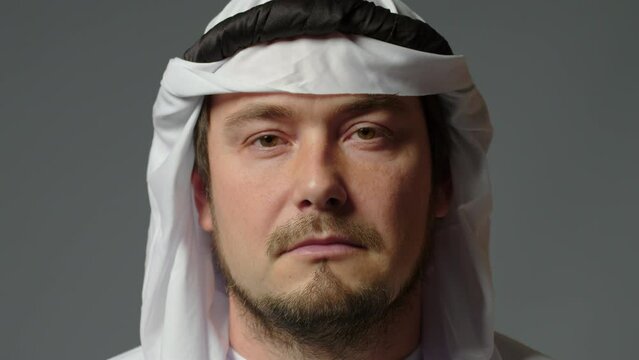 The sheikh wearing a white kandura and a gutra on his head close up looks at camera gray background