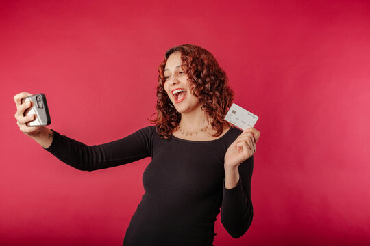 Cute redhead woman wearing black dress standing isolated over red background taking a selfie with credit card and looks very happy. Make selfie by phone holding credit card.