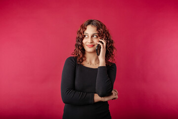 Woman wearing black ribbed dress standing isolated over red background looks at the empty copy space out of the corner of eye while talking on the phone. Discusses something interesting with friend.