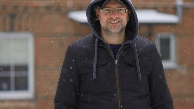 Smiling Happy Man In Hoodie Inside Falling Snowflakes In Front Of House During Heavy Snowfall Slow Motion. Snowstorm And Blizzard In Winter Season