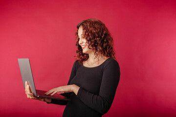 Redhead woman standing isolated over red background holding a laptop with a confident expression....