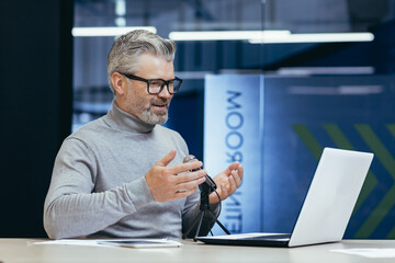 Mature smiling businessman recording audio podcast about finance and strategy to achieve all and good achievement, gray haired man inside office using professional microphone and laptop.