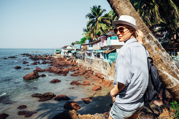 A beautiful tourist girl in dark glasses, a white hat and a blue shirt looks at the sea, palm trees and coastal houses. A concept for travel and tourism
