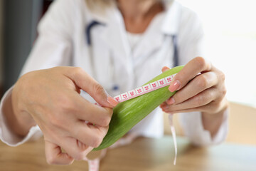 Doctor measures length of cucumber with measuring tape while sitting at table in medical clinic. - obrazy, fototapety, plakaty