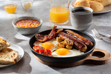 Full English breakfast with fried eggs, sausages, beans, bacon, grilled tomatoes, mushrooms, toasts and cup of coffee on gray background.