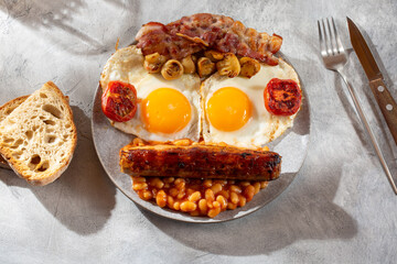 Full English breakfast with fried eggs, sausages, beans, bacon, grilled tomatoes, mushrooms, toasts and cup of coffee on gray background.