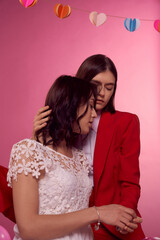 Valentine's day concept. Studio portrait of two young lesbian girls. One girl is in a white dress, the second woman is in a red suit. They are sitting on a pink background. Cropped shot.