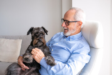 Happy Grandpa is sitting at chair in home with dog Jack Russell Terrier on his legs. Senior man...