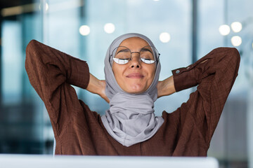 Portrait of a young businesswoman in hijab and glasses sitting in the office at the table in front of the computer. She put her hands behind head, closed her eyes, resting, taking a break, smiling.